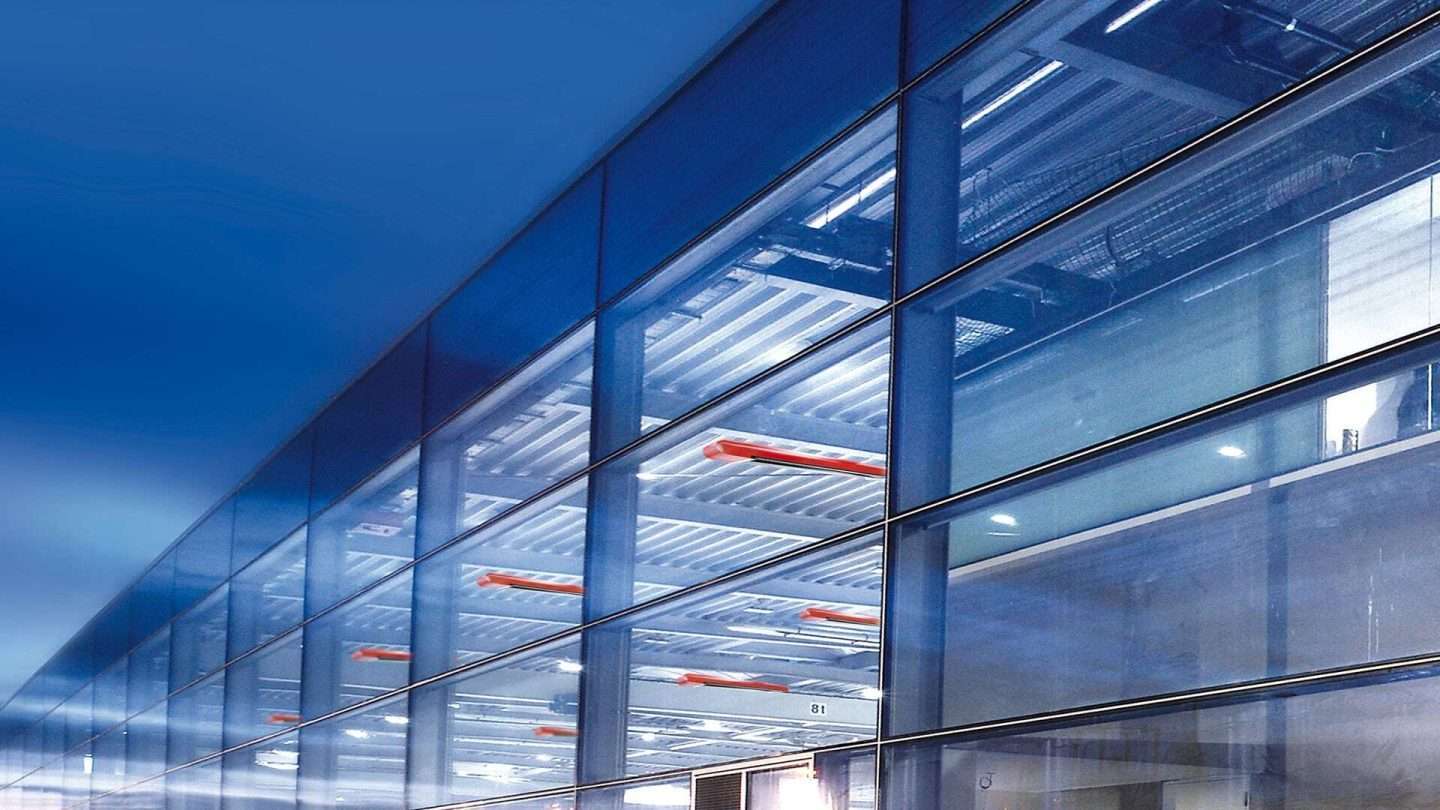 Exterior view of a large hall illuminated from the inside. The cold blue light makes the appealing design of the red MAXIMA dark spotlights stand out.