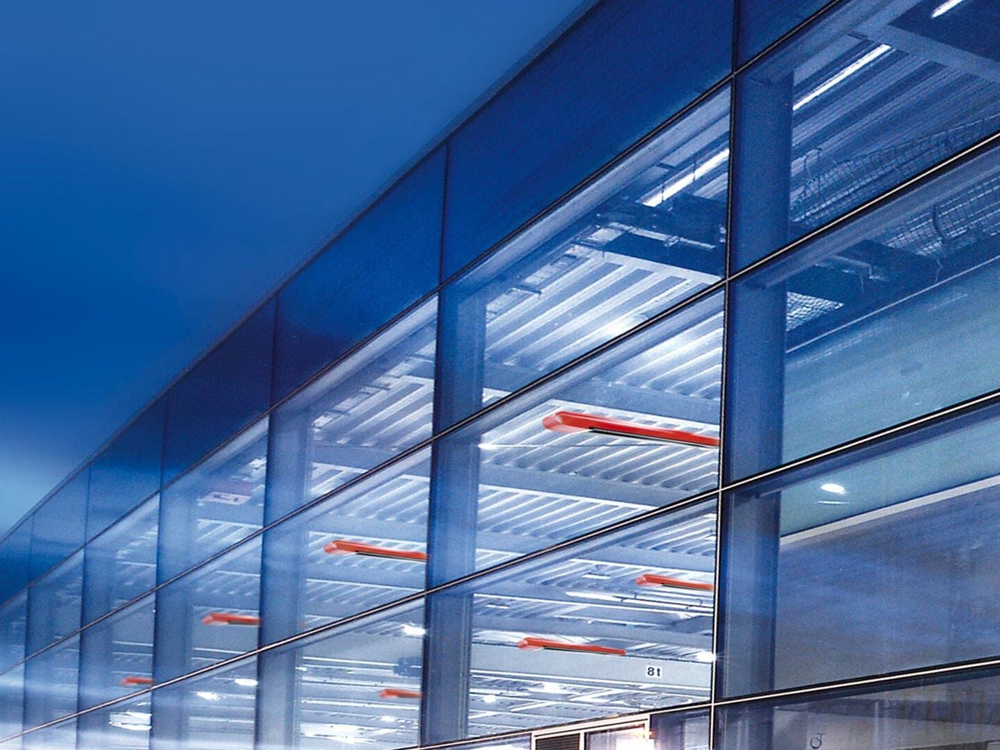 Außenansicht einer großen von innen beleuchteten Halle. Das kalte blaue Licht, lässt ansprechende Design der roten MAXIMA Dunkelstrahler hervorstechen.