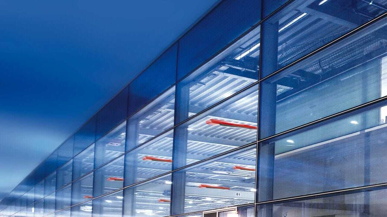 Außenansicht einer großen von innen beleuchteten Halle. Das kalte blaue Licht, lässt ansprechende Design der roten MAXIMA Dunkelstrahler hervorstechen.